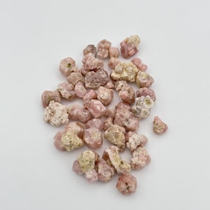 Pode incluir: Um monte de pequenos cristais brutos, naturais, não polidos, de cor rosa e branca. Os cristais são de várias formas e tamanhos.