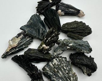 May include: A collection of raw black tourmaline crystal specimens featuring striated, elongated formations with jagged edges and occasional white mineral inclusions. These dark, opaque stones exhibit a natural, fibrous texture and varying geometric shapes.