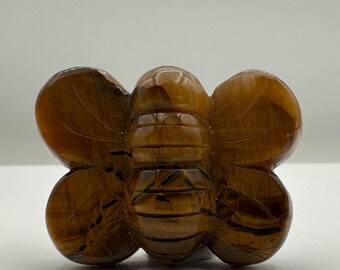 May include: A polished tiger's eye gemstone carved into the shape of a bee. The carving features a rounded head, a segmented body, and two symmetrical wings on each side, displaying the natural golden-brown chatoyant bands of the stone.