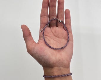 May include: Two amethyst beaded bracelets. One bracelet is on a wrist, and the other is held in an open hand. The bracelets have small, round, purple beads and silver-colored clasps. The background is a plain white.