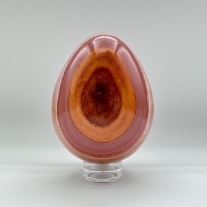 Può includere: Una pietra lucidata a forma di uovo con bande concentriche rosa, arancio e marrone. La pietra è esposta su un supporto in acrilico trasparente.