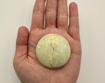 May include: A smooth, circular, pale yellow stone with a speckled texture and a prominent, jagged gold-colored vein running vertically through the center. The object is round and flat, featuring a polished surface with natural-looking variations.