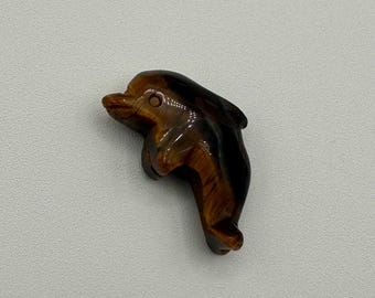 May include: A small, hand-carved dolphin figurine crafted from polished tiger's eye gemstone. The stone features a natural chatoyant pattern with alternating bands of golden-brown and dark brown colors, highlighting the dolphin's curved body shape.