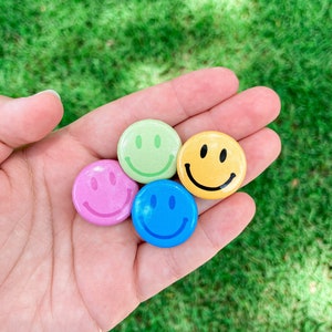May include: Four smiley face pins in different colors: green, yellow, pink, and blue. The pins are held in a hand against a green grassy background.