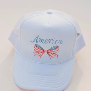 May include: White trucker hat with a red, white, and blue bow and the word "America" printed on the front.