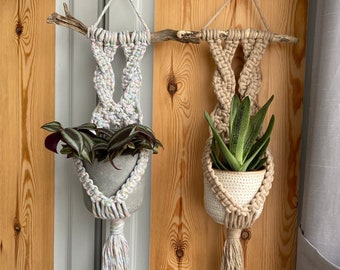 May include: Two macrame plant hangers with wooden dowels and tassels. The left hanger is white with rainbow colored tassels and holds a green plant. The right hanger is beige with beige tassels and holds a green succulent.