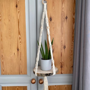 May include: A white macrame plant hanger with a circular wooden shelf supporting a potted plant with green leaves. The hanger has wooden beads and a tassel. The background shows wooden doors and a grey curtain.