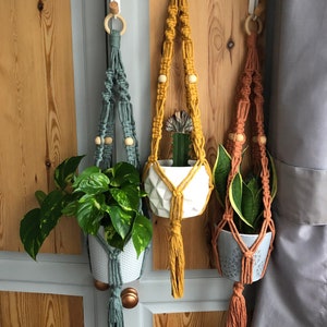 May include: Three macrame plant hangers in teal, mustard yellow, and terracotta, each holding a potted plant. The hangers feature wooden beads and tassels, suspended from wooden rings. The plants include a pothos, a cactus, and a snake plant.