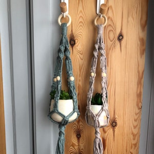 May include: Two macrame plant hangers, one teal and one beige, each holding a small white ceramic pot with a green plant. The hangers have wooden beads and tassels, suspended from wooden rings and hooks.