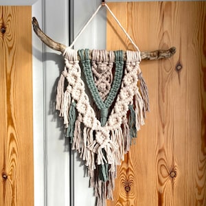 May include: A macrame wall hanging with a natural wood branch, featuring a geometric design in shades of beige, cream, and sage green.