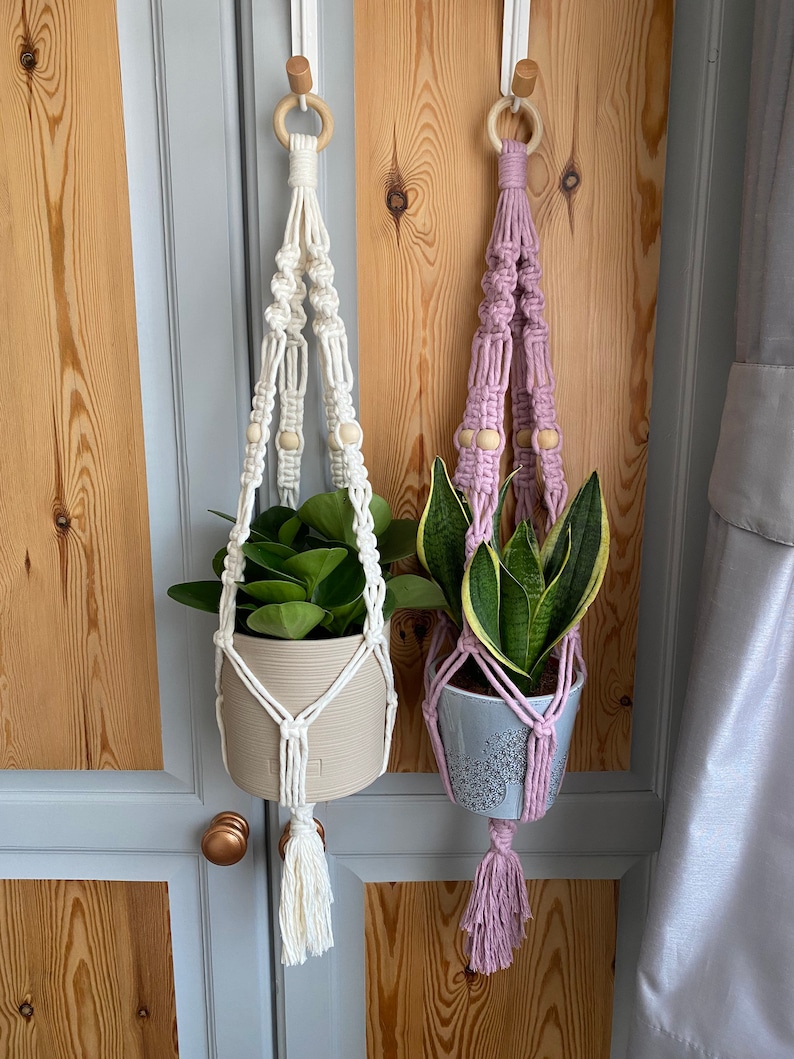 May include: Two macrame plant hangers, one white and one lavender, each holding a potted plant. The white hanger has a beige pot with green leaves, while the lavender one has a gray pot with a green and yellow plant. Both are suspended from wooden rings.