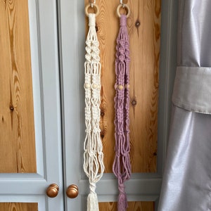 May include: Two macrame plant hangers, one cream and one lavender, hang from wooden rings. Each has wooden beads and a tassel. The hangers are suspended against a light wood door with gray trim and a gray curtain.