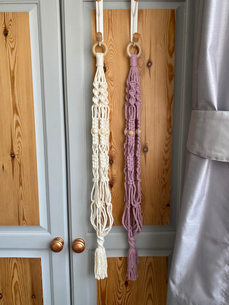 May include: Two macrame plant hangers, one cream and one lavender, hang from wooden rings. Each has wooden beads and a tassel. The hangers are suspended against a light wood door with gray trim and a gray curtain.