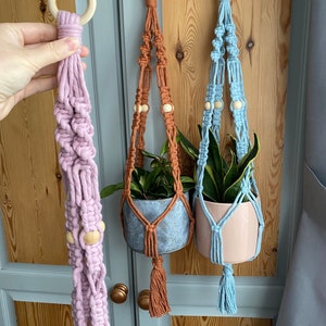 May include: Three macrame plant hangers in shades of lavender, rust, and light blue. Each hanger features a wooden ring at the top, wooden beads, and a tassel. The hangers are designed to hold small potted plants, adding a touch of bohemian style.