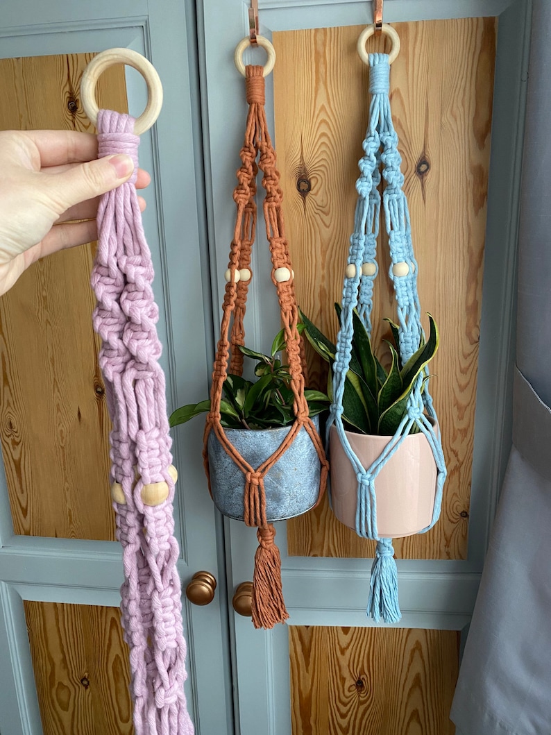 May include: Three macrame plant hangers in shades of lavender, rust, and light blue. Each hanger features a wooden ring at the top, wooden beads, and a tassel. The hangers are designed to hold small potted plants, adding a touch of bohemian style.