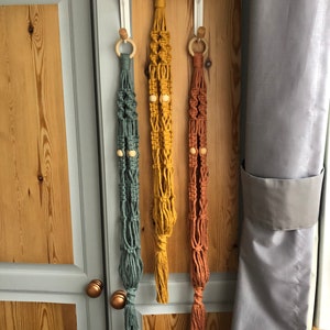 May include: Three macrame plant hangers in teal, mustard yellow, and rust colors. Each hanger features a wooden ring at the top and wooden beads. The hangers are suspended from white over-the-door hooks, showcasing a bohemian style.