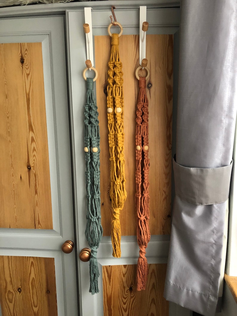 May include: Three macrame plant hangers in teal, mustard yellow, and rust colors. Each hanger features a wooden ring at the top and wooden beads. The hangers are suspended from white over-the-door hooks, showcasing a bohemian style.