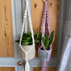 May include: Two macrame plant hangers, one white and one lavender, each holding a potted plant. The white hanger has a beige pot with green leaves, while the lavender one has a gray pot with a green and yellow plant. Both are suspended from wooden rings.