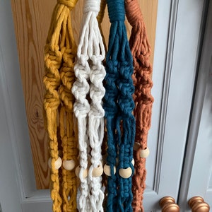 May include: A collection of five macrame plant hangers in various colors: mustard yellow, white, teal, and rust. Each hanger features a wooden ring at the top and wooden beads. The hangers are displayed against a neutral background.
