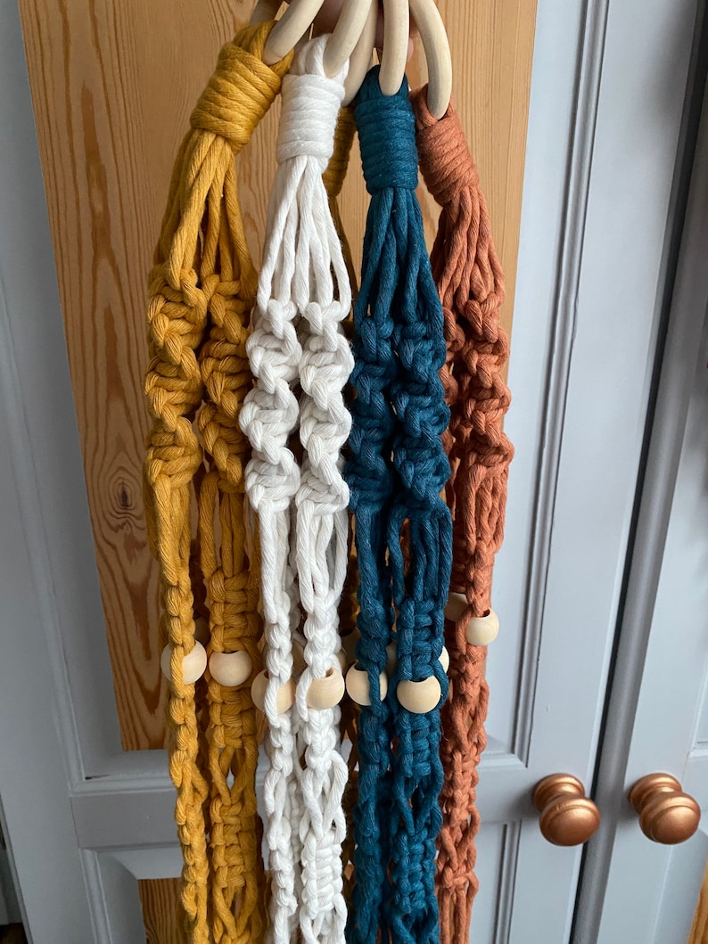 May include: A collection of five macrame plant hangers in various colors: mustard yellow, white, teal, and rust. Each hanger features a wooden ring at the top and wooden beads. The hangers are displayed against a neutral background.
