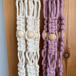 May include: Close-up of two macrame plant hangers. One is off-white, the other is a soft lavender. Both feature wooden beads and intricate knotting. The hangers are displayed against a wooden background, showcasing the texture and craftsmanship.