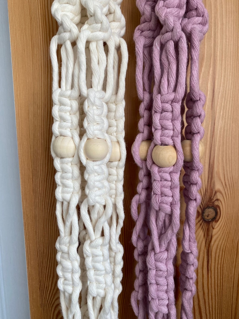 May include: Close-up of two macrame plant hangers. One is off-white, the other is a soft lavender. Both feature wooden beads and intricate knotting. The hangers are displayed against a wooden background, showcasing the texture and craftsmanship.