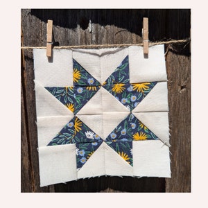 Puede incluir: Un bloque de colcha de tela blanca con un estampado floral azul marino y amarillo. El bloque tiene forma de estrella y está colgado de dos pinzas de madera.