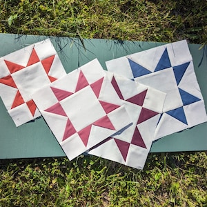 May include: Four fabric squares with a star design. The squares are in red, blue, and two shades of pink. The squares are arranged on a green surface.