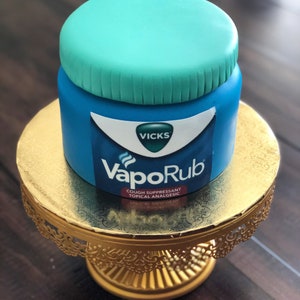 Könnte beinhalten: Ein blauer und grüner Kuchen, der wie ein Vicks VapoRub-Glas dekoriert ist. Der Kuchen steht auf einem goldenen Kuchenständer.