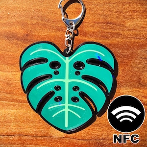 May include: A teal and black monstera leaf-shaped keychain with a silver keyring. The keychain has a detailed leaf design with a black outline and a light green center. A black and white NFC symbol is on the bottom right.