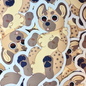 May include: A pile of stickers depicting a cartoon hyena. The hyena is tan with brown spots, big eyes, and a pink tongue. Each sticker has a white border, creating a clean outline.