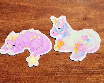 May include: Two colorful cartoon stickers, one of a purple and yellow dinosaur sleeping and the other of a white unicorn with a pink mane and tail. Both stickers have a sparkly, iridescent finish.