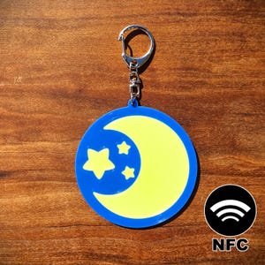 May include: A blue and yellow keychain featuring a crescent moon and stars design. The keychain has a silver clasp and a black NFC logo with the text "NFC". The design is on a circular pendant.