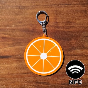 May include: Orange slice keychain with a silver keyring. The keychain features a bright orange design with white segments, resembling a citrus fruit. The keychain also has an NFC symbol.