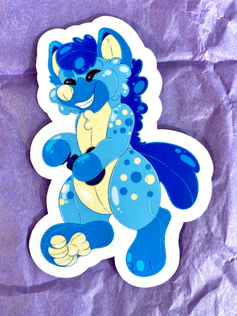 May include: A blue and yellow plush toy with a smiling face and white spots. The toy is standing on its hind legs and holding a black controller in its front paws.