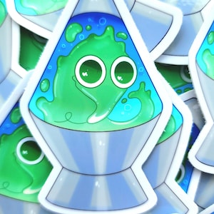 Puede incluir: Una pegatina con un diseño de lámpara de lava de dibujos animados. La lámpara tiene una combinación de colores azul y verde, con una mancha verde sonriente en el interior. La base es plateada y la pegatina tiene un borde blanco.