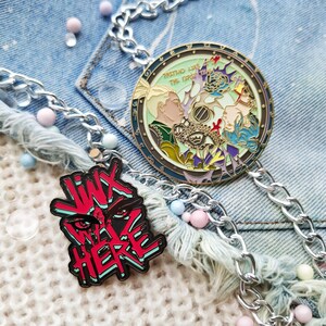 May include: Two enamel pins on a silver chain. The first pin is round and features a colorful illustration of two people with the text "Pretend Like The Dream Never Died". The second pin is red and black and features the text "Wix We're Here" with a stylized eye.