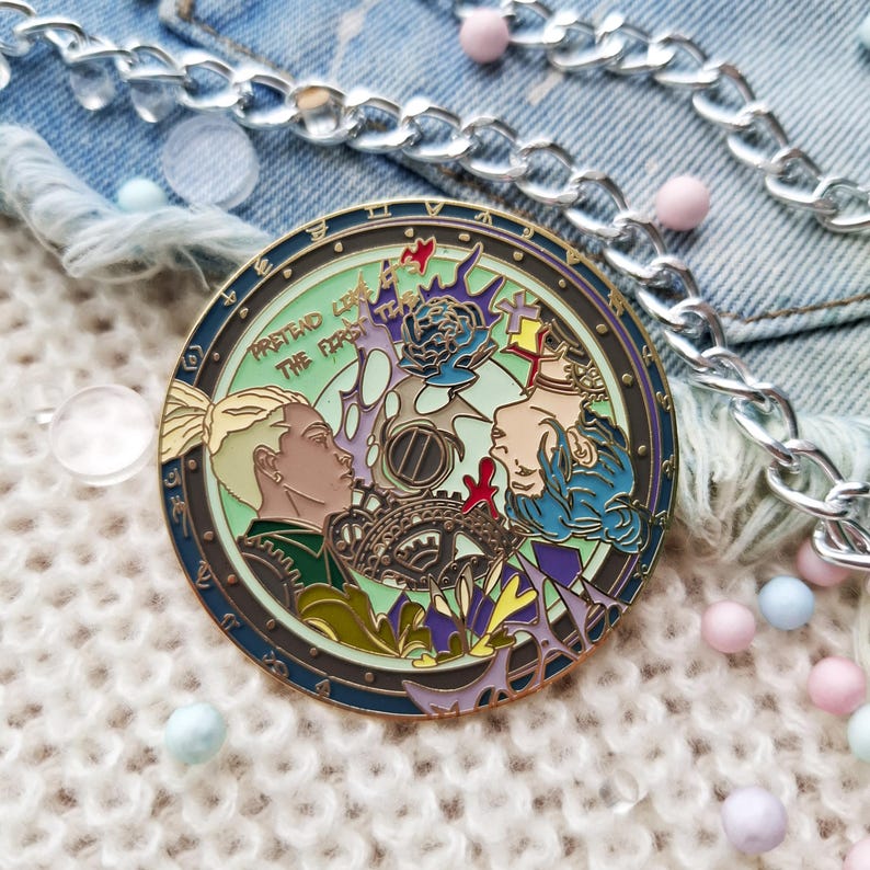 May include: A gold-toned enamel pin featuring two figures with blue and green hair. The pin is circular and has a green background with a floral design. The text "Pretend Like The Fire Is The First Time" is written around the edge of the pin.
