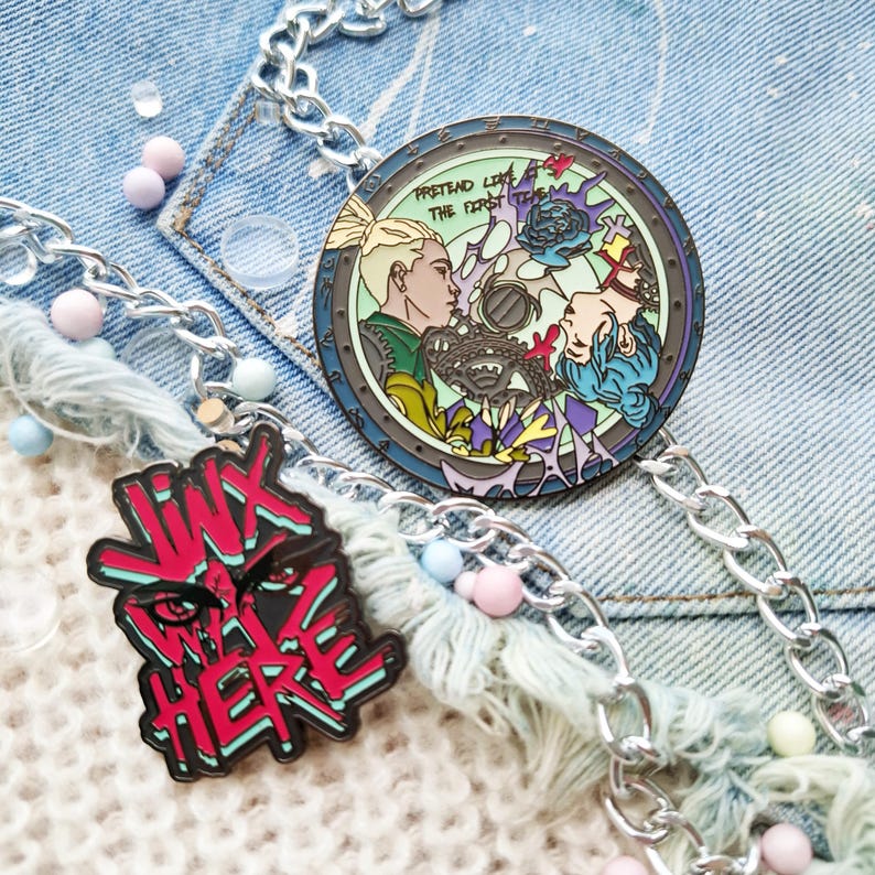 May include: Two enamel pins on a denim background. One pin is round and features a colorful illustration of two people with the text "Pretend Like The First Time". The other pin is red and black and features the text "I'm Not Here".
