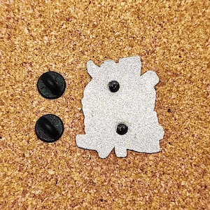 May include: The back of a silver enamel pin with a detailed design. The pin has two black rubber stoppers on the back.