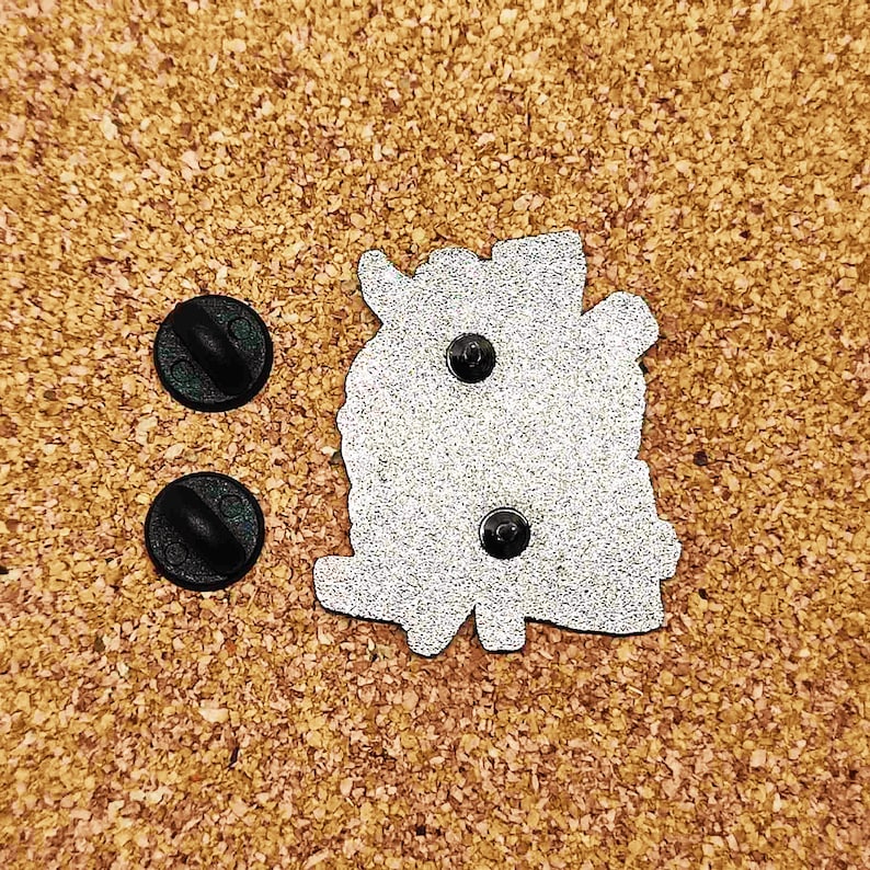 May include: The back of a silver enamel pin with a detailed design. The pin has two black rubber stoppers on the back.