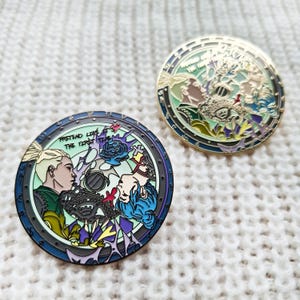 May include: Two round enamel pins with a gold finish. The first pin features a colorful illustration of two people with the text "Pretend Like The First Time". The second pin features a colorful illustration of a circle with a floral design.