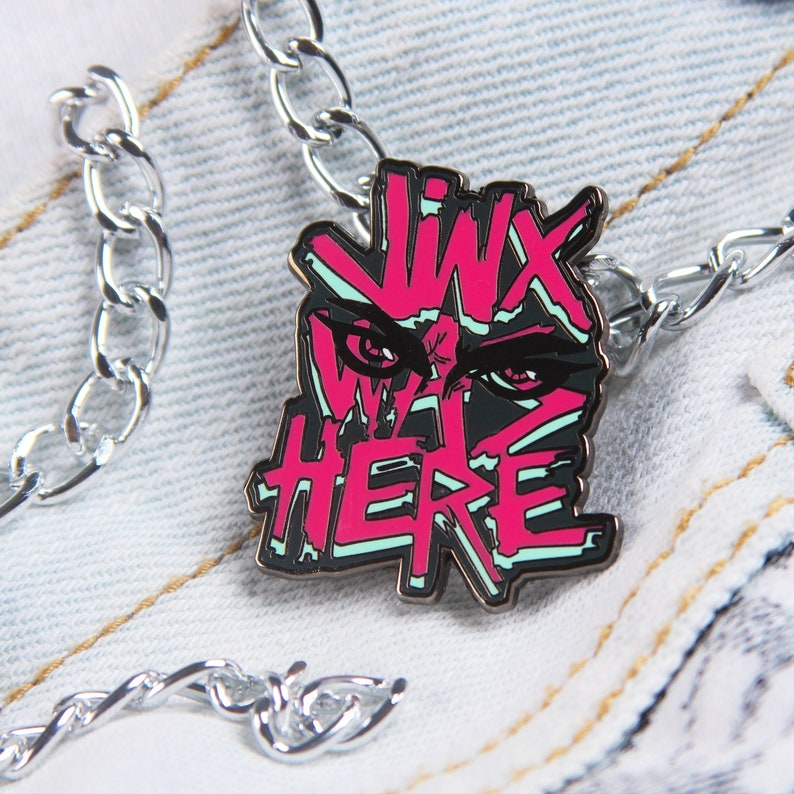 May include: A silver chain necklace with a pink and black enamel pin featuring a stylized eye and the text "Jinx Was Here".
