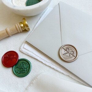 May include: A white marble bowl filled with various wax seals, including red, green, and gold seals with different designs. A gold and wooden wax seal stamp is on the table next to the bowl. A silver wax seal with a bow design is on a gray envelope.