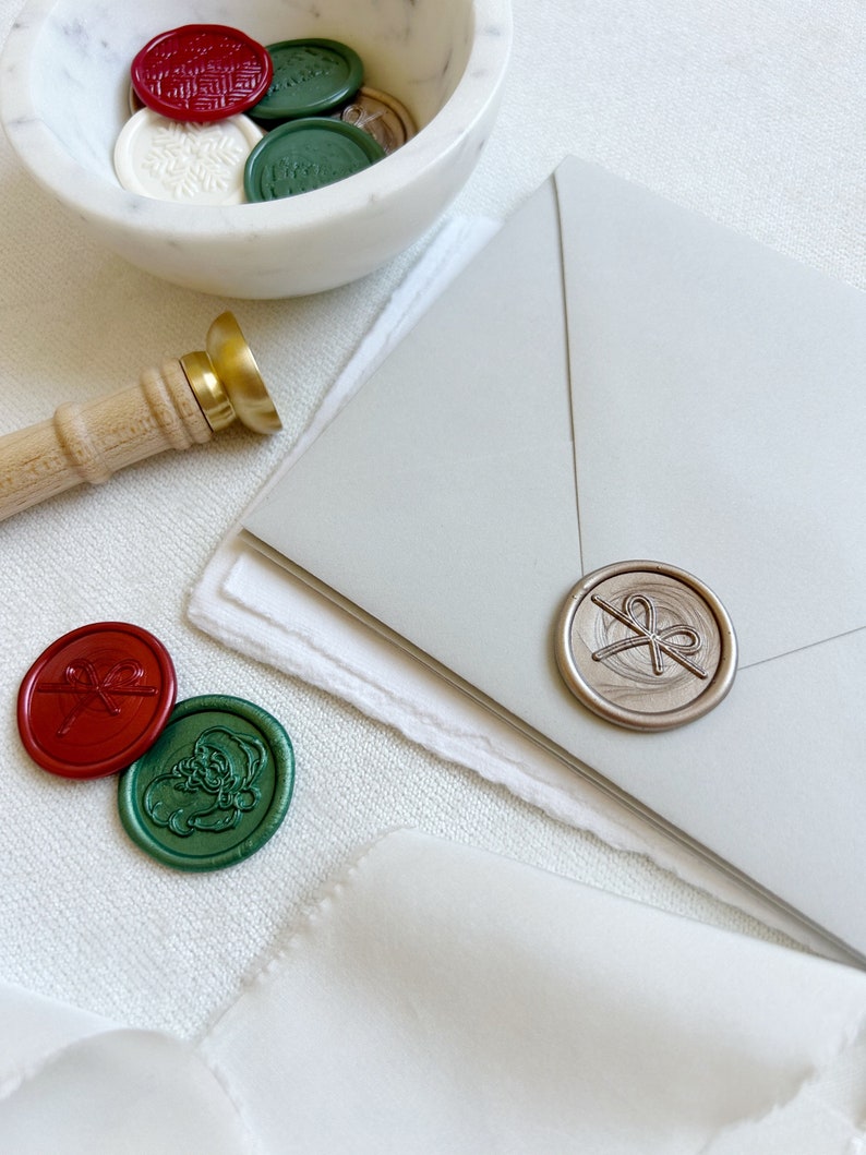 May include: A white marble bowl filled with various wax seals, including red, green, and gold seals with different designs. A gold and wooden wax seal stamp is on the table next to the bowl. A silver wax seal with a bow design is on a gray envelope.