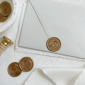May include: Gold wax seals with a floral design and the letter "S" in the center. The seals are on a white surface with a white envelope and a white ribbon.