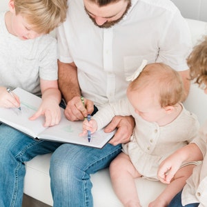 May include: A father and two children are sitting on a white couch, coloring in a book with crayons. The father is wearing a white shirt and blue jeans. The older child is wearing a gray shirt and blue jeans. The younger child is wearing a white shirt and has a white bow in their hair.