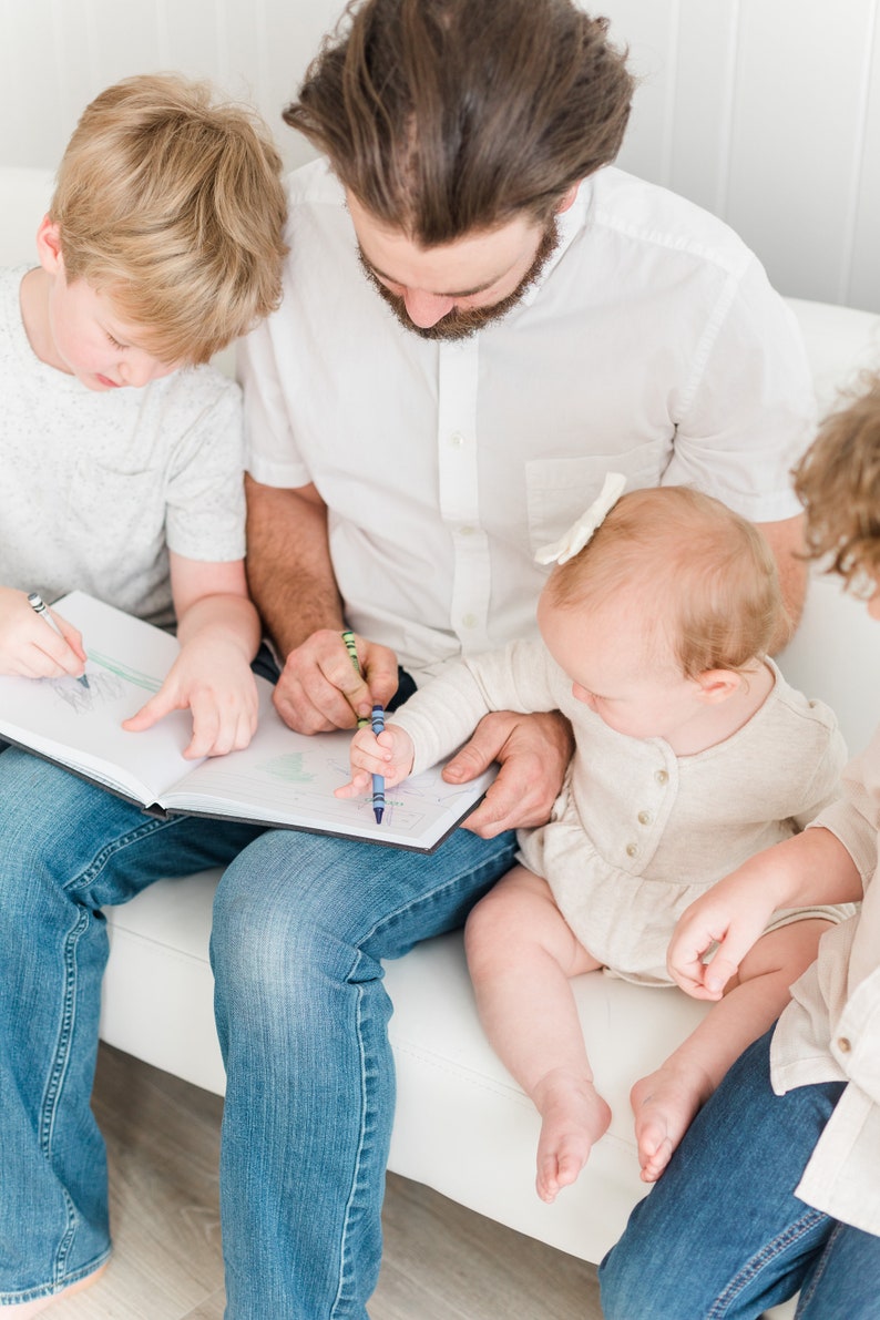 May include: A father and two children are sitting on a white couch, coloring in a book with crayons. The father is wearing a white shirt and blue jeans. The older child is wearing a gray shirt and blue jeans. The younger child is wearing a white shirt and has a white bow in their hair.