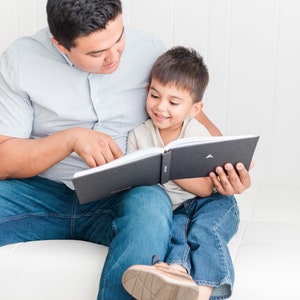 May include: A father and son sit on a white couch, reading a black hardcover book together. The father is wearing a light blue button-down shirt and blue jeans. The son is wearing a light beige shirt and blue jeans.