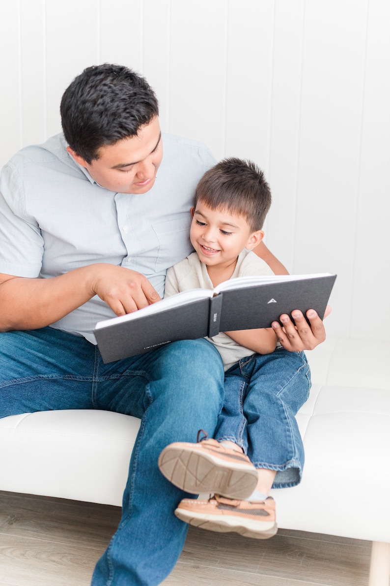 May include: A father and son sit on a white couch, reading a black hardcover book together. The father is wearing a light blue button-down shirt and blue jeans. The son is wearing a light beige shirt and blue jeans.